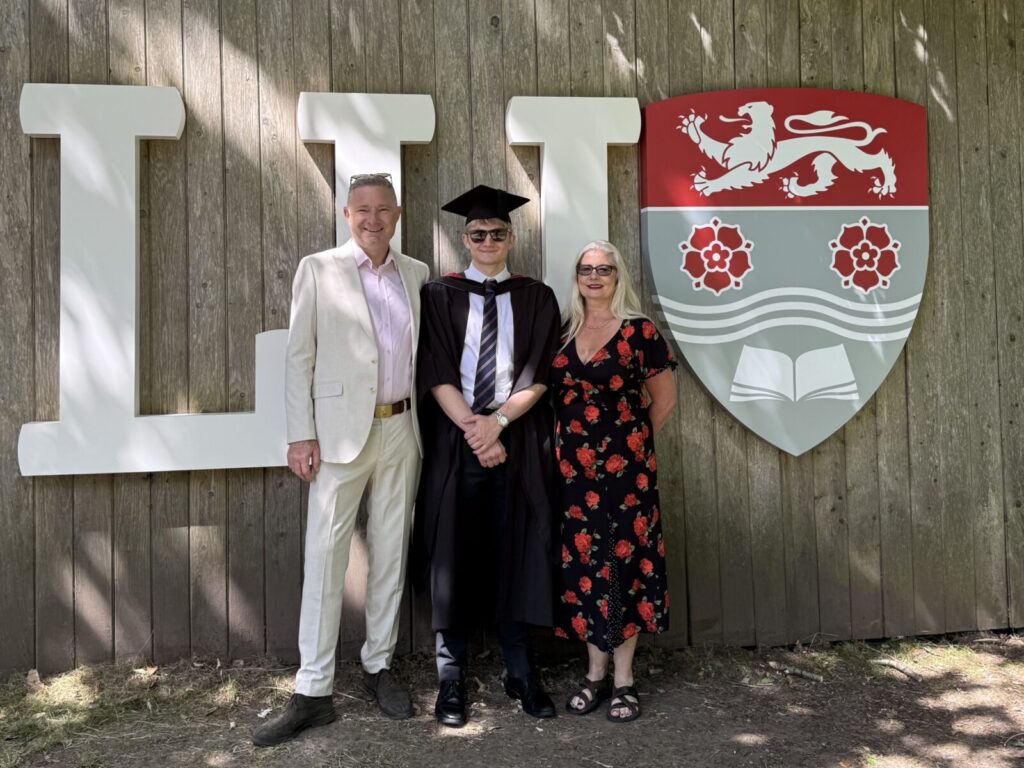 Graduate with two adults smiling by university crest, Leeds; celebrating academic achievement outdoors.